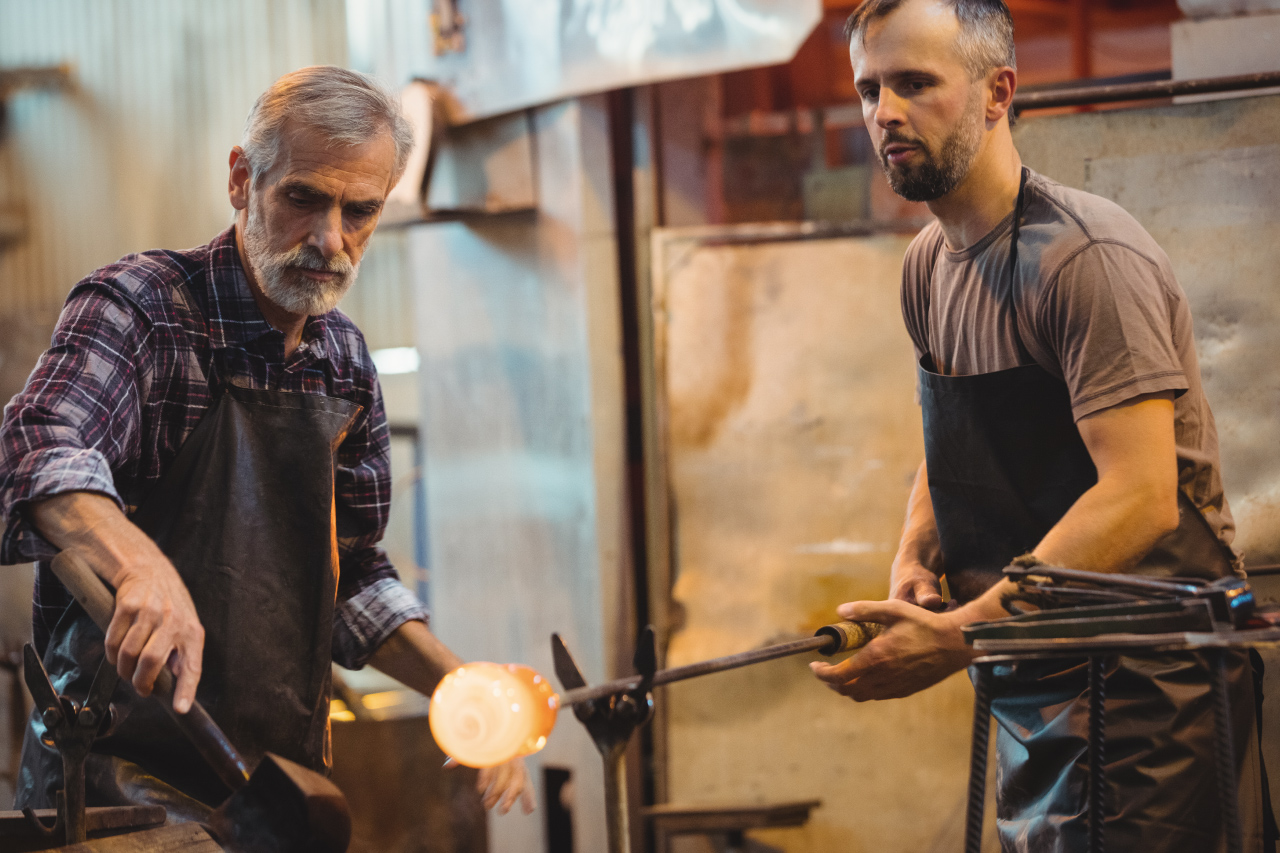 Dois homens, trabalham na produção de vidros, um deles segura a peça incandescente.