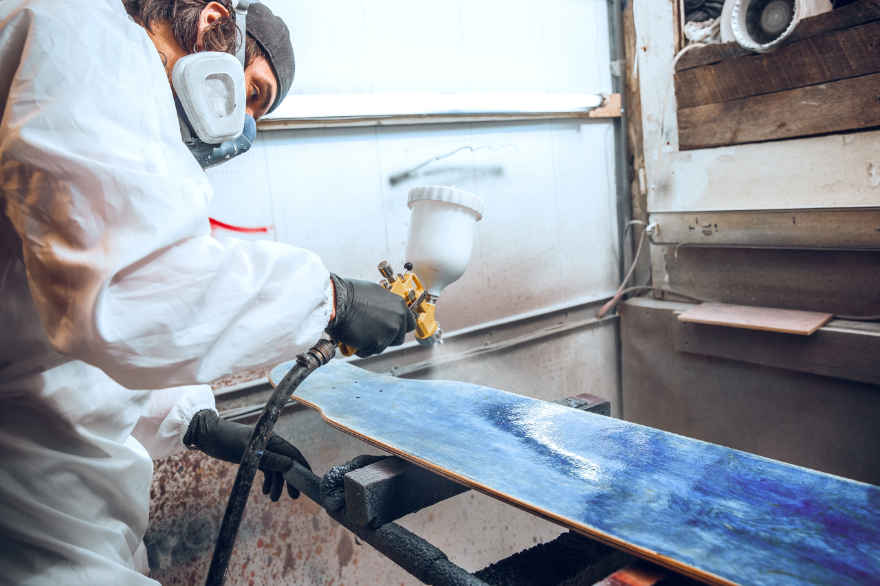 Um homem numa cabine de pintura, segura uma pulverizadora e aplica tinta azul sobre uma peça de madeira.