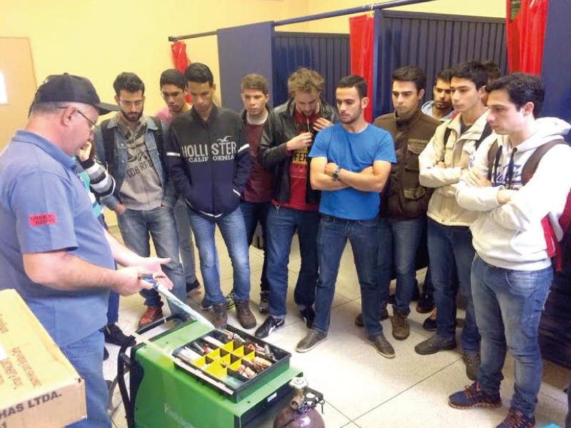 Dez jovens lado a lado observam um homem que mostra o equipamento do carrinho de solda mig.