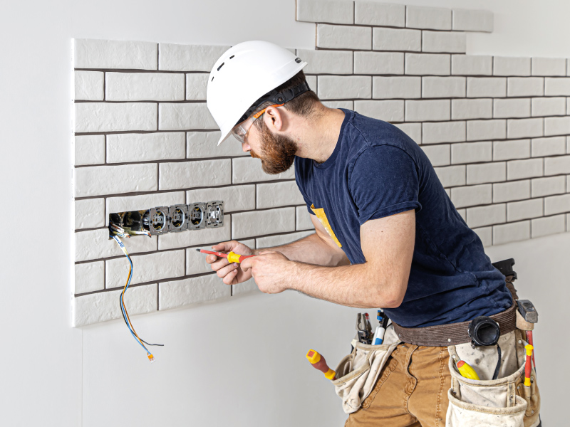 Um homem de capacete branco, óculos de proteção e cinto para ferramentas segura uma chave próxima de uma caixa de tomadas de energia embutidas. 